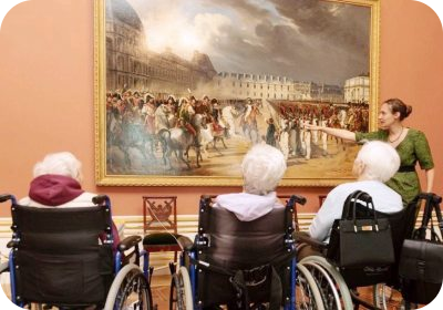 Молодая женщина-экскурсовод в Эрмитаже показывает на картину пожилым людям в инвалидных колясках